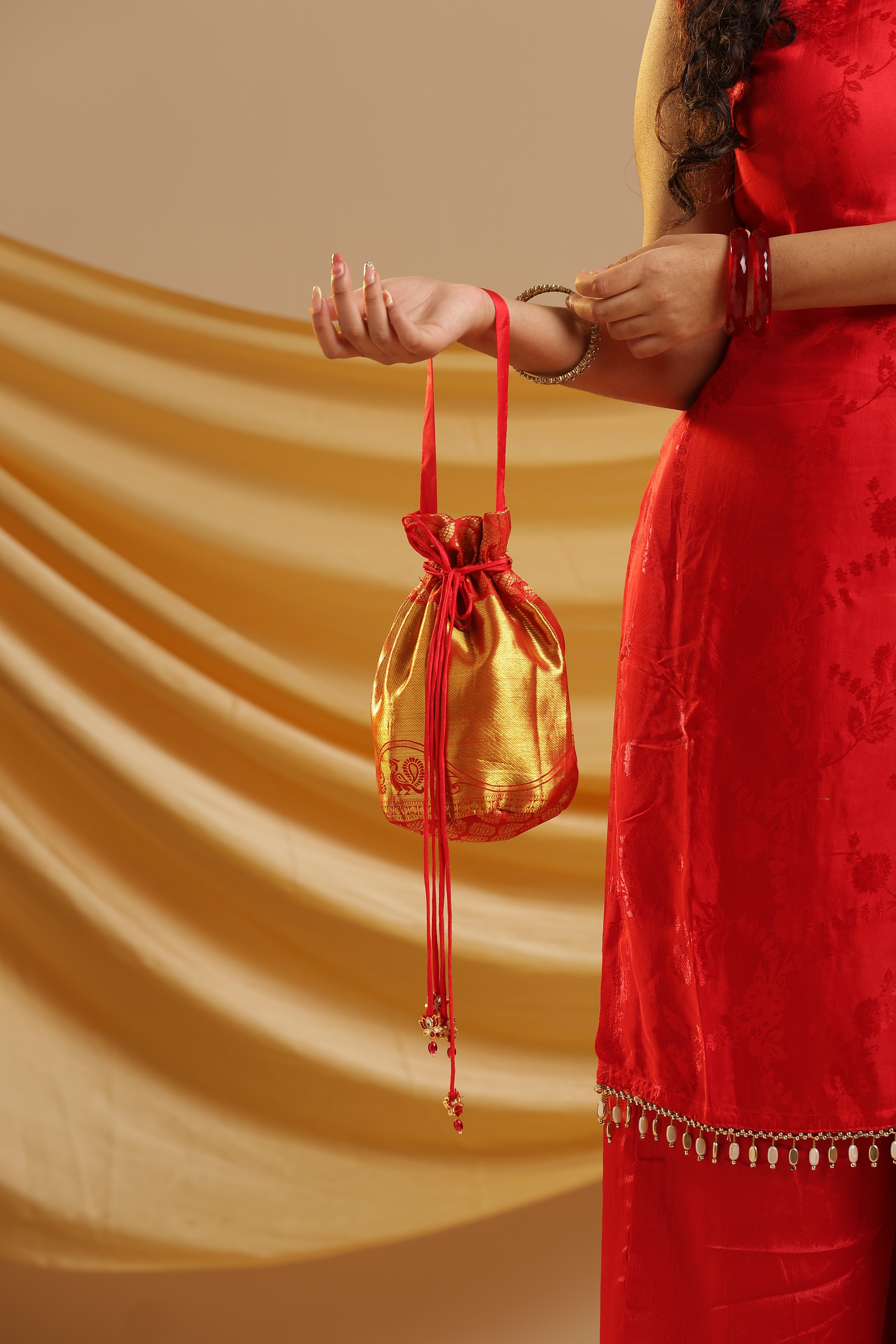 A woman in a red traditional dress holds a small shiny gold drawstring pouch with red ties against a draped gold fabric background.