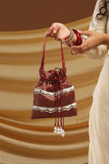 A woman in a cream-colored kurta holds a small maroon drawstring pouch decorated with white lace and bead tassels, set against a draped brown background.