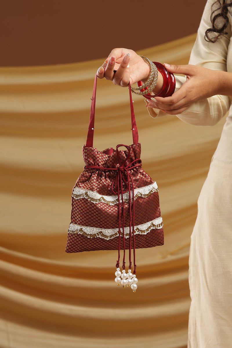 A woman in a cream-colored kurta holds a small maroon drawstring pouch decorated with white lace and bead tassels, set against a draped brown background.