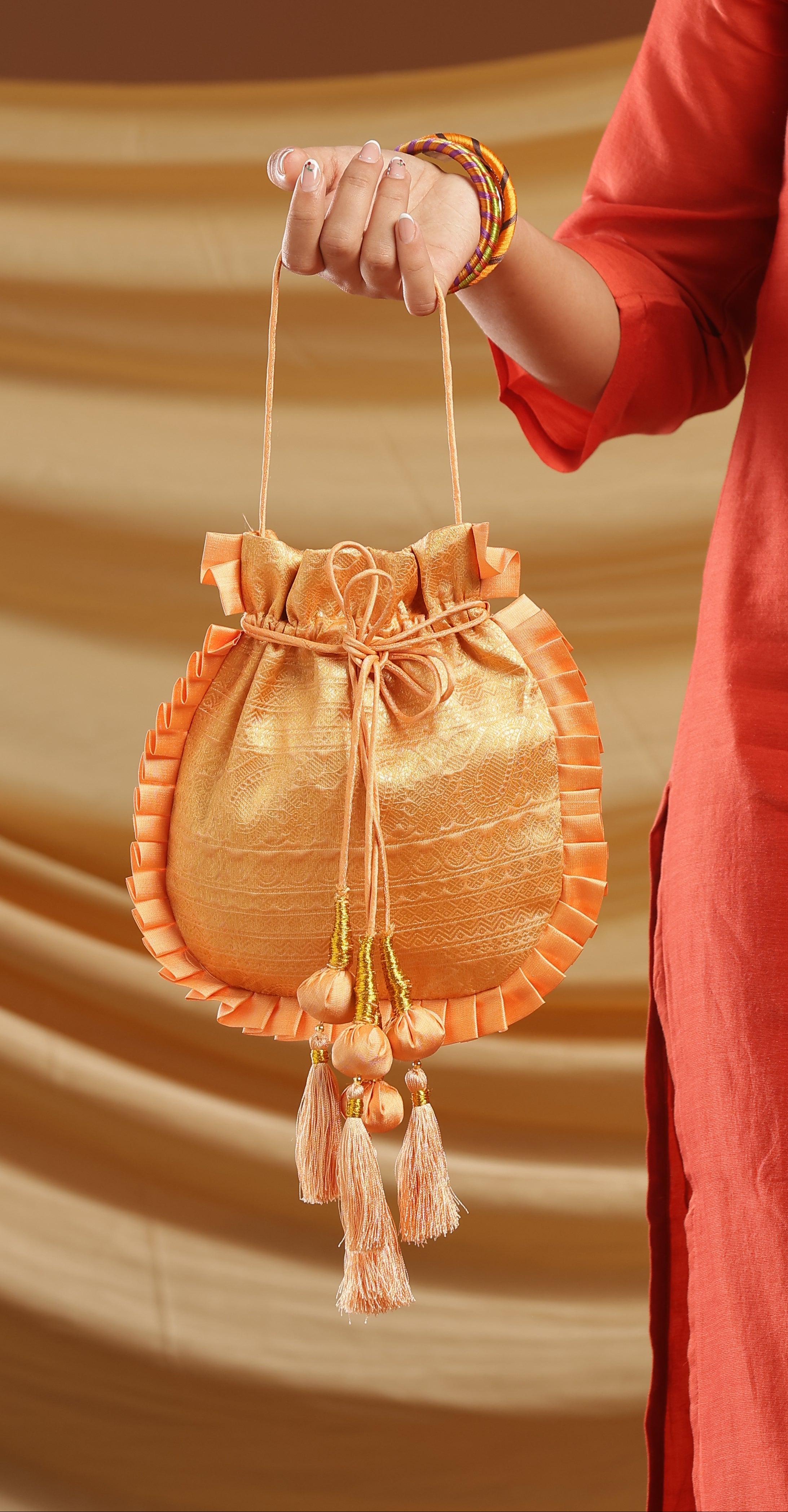 A woman wearing a rust-orange embroidered kurta holds a small metallic gold drawstring pouch with tassels against a warm brown studio backdrop.