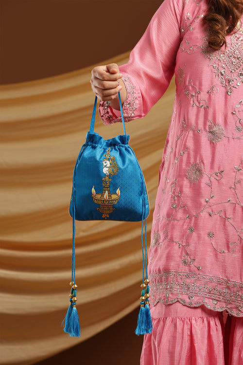 A person in a pink embroidered traditional outfit holds a small blue velvet drawstring pouch with gold embroidery and tassels against a warm brown draped backdrop.