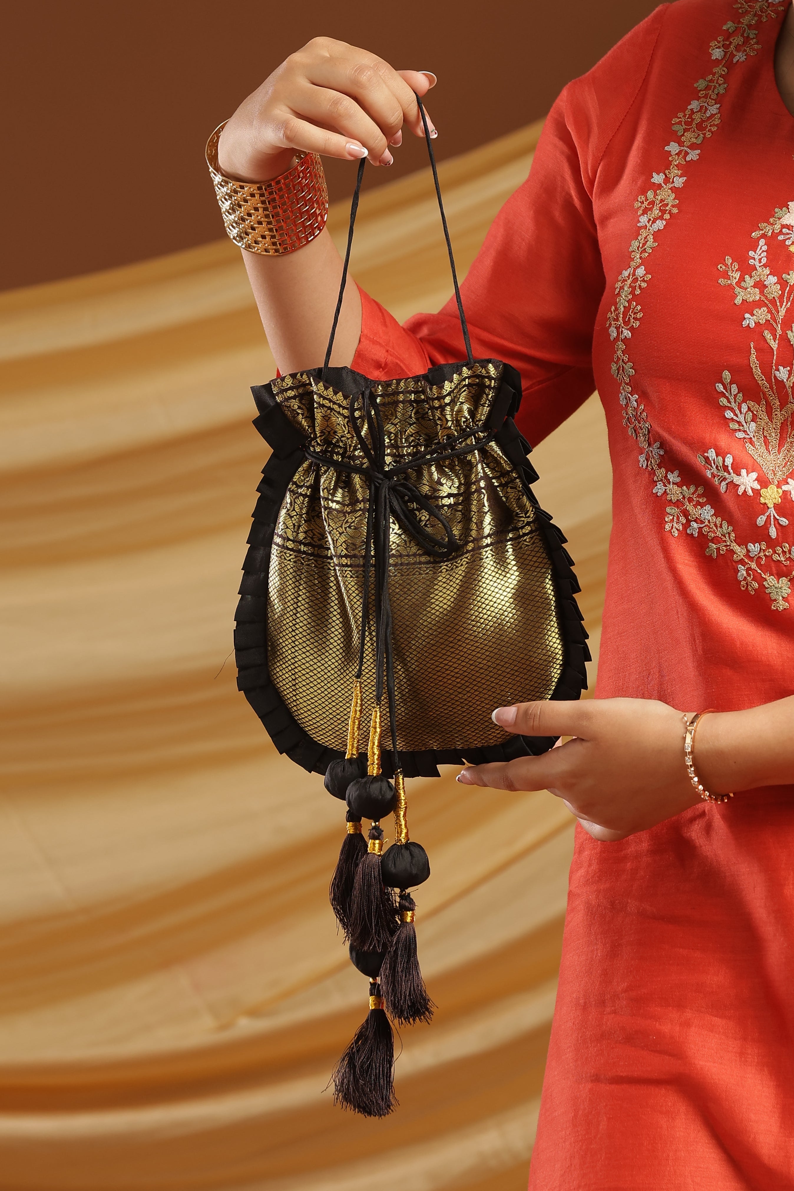 A woman in a red embroidered kurta holds up a small black and gold drawstring pouch bag against a warm brown studio background.