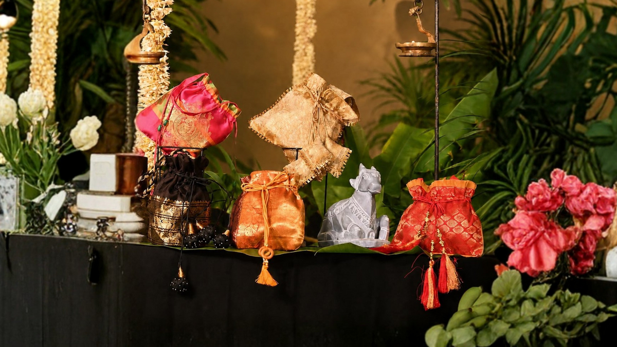 A decorative altar-like display with hanging garlands, warm string lights, green plants, flowers, small figurines, and orange fabric bundles arranged against a warm brown wall.