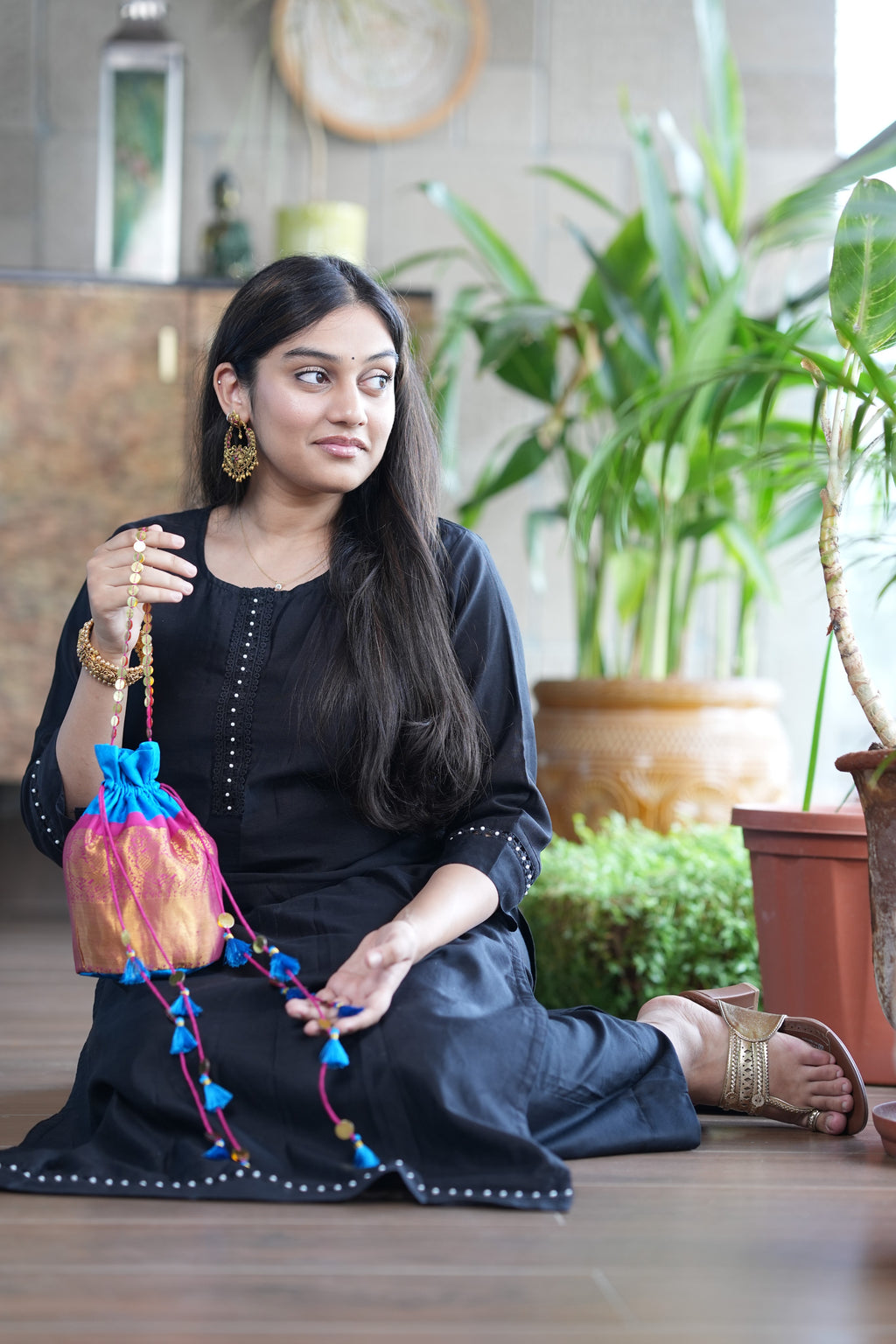 Woman in a black traditional outfit holding a colorful bag in a home setting with plants.