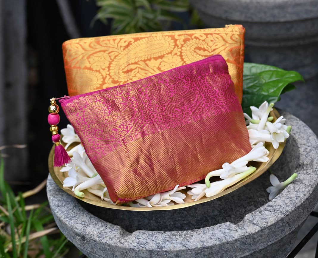 Two ornate silk pouches or small bags made from patterned zari fabric, one in orange-gold and one in magenta-pink, displayed on a brass tray with white flowers on a stone pedestal in a garden setting.