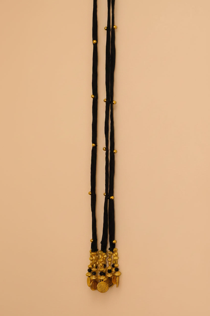 A long black velvet necklace with gold bead accents and a gold-toned coin pendant, hanging on a beige wall next to a white door frame.